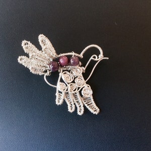 Pode incluir: Um broche de pássaro de fio de prata com um acento de pedra preciosa roxa. O pássaro tem um bico longo e fino e um design de asa detalhado.
