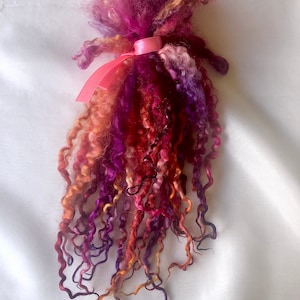 May include: A handmade fiber art doll with a pink ribbon. The doll features a mix of curly, colorful wool fibers in shades of purple, pink, orange, and red. The doll's form is abstract, with a fluffy head and flowing, textured strands.