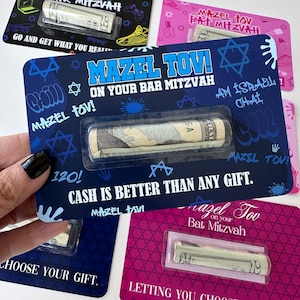 May include: A blue and white card with a Star of David design and the text "Mazel Tov! On Your Bar Mitzvah" and "Cash is better than any gift." A rolled up banknote is visible in a clear plastic pocket on the card.