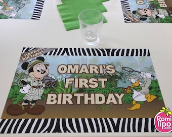 May include: A birthday party table setting featuring placemats with Mickey Mouse and Donald Duck in safari attire. The placemats have a zebra stripe border and text that reads "Omari's First Birthday". A clear glass and green napkins are also on the table.