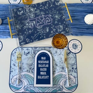 May include: A Passover Seder table setting with a blue floral design. The set includes a serving tray with gold handles, a placemat with the text "Mah Nishtanah Halaylah Haze Mikol Halaylot?", a glass, and a small round coaster.