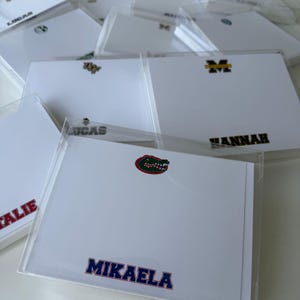 May include: White notepads with personalized names and logos, encased in clear plastic. The notepads feature names like "Lucas," "Hannah," and "Mikaela" in various fonts and colors. Some notepads have university logos, including a Gator logo.