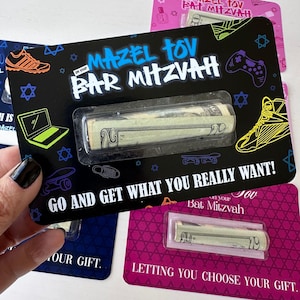 May include: Black card with the text "Mazel Tov on your Bar Mitzvah" and a rolled up banknote inside a plastic sleeve. The card also has a skateboard, a video game controller, and a sneaker graphic. The text "Go and get what you really want!" is printed on the card.