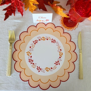 May include: A round scalloped paper placemat featuring a central wreath of autumn leaves and berries in shades of orange, red, and yellow. A white place card with the name Olivia and a matching leaf motif sits above the placemat.
