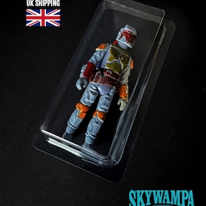 May include: A Boba Fett action figure encased in a clear plastic blister pack. The figure features a light gray jumpsuit, a green chest plate, orange shoulder and knee armor, and a helmet with a red visor. The text SKYWAMPA and WWW.SKYWAMPA.COM appear.