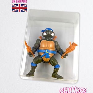 May include: A Teenage Mutant Ninja Turtle action figure in a clear plastic case. The figure is green, with a blue mask, orange weapons, and a brown shell. The figure is in a fighting stance.
