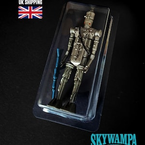 May include: A silver-colored droid action figure with a slender, mechanical frame and a cylindrical head, encased in a clear plastic blister pack. The figure holds a long, blue rifle-like accessory in its right hand and features intricate molded details.