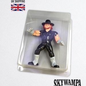 May include: A plastic action figure of a wrestler wearing a black hat, a black sleeveless shirt with tattered edges, black pants, and white boots with grey gloves, encased in a clear rectangular plastic blister pack packaging.