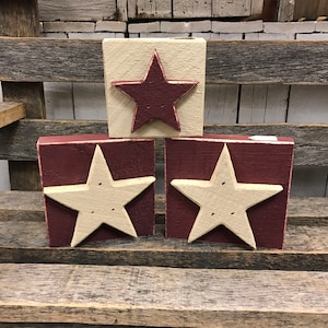 May include: Three wooden blocks with a red and white painted star design. The stars are painted in a distressed style with a white star on a red background and a red star on a white background.