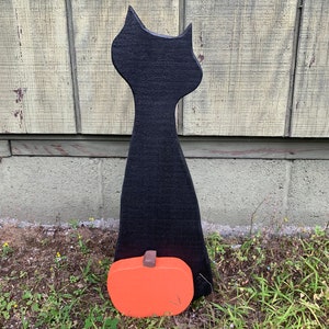 May include: Black wooden cat silhouette with a small orange pumpkin at its feet. The cat is standing on a patch of green grass.