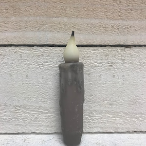 May include: A brown candle with a white wax tip and a black wick. The candle is sitting on a white wooden surface.