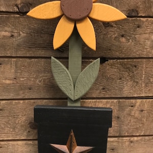 May include: A wooden sunflower with a brown center and yellow petals is in a black pot with a gold star. The pot is on a black base.