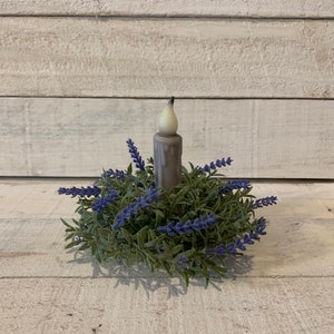 May include: A small, white candle with a gray base sits in a wreath of purple lavender and green leaves.