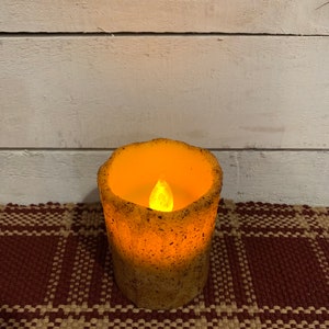 May include: A small, round, beige candle with a flickering flame inside. The candle is sitting on a brown and beige plaid surface.