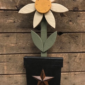 May include: A wooden daisy flower with a yellow center, green leaves, and a black painted pot with a rusty star. The flower is standing on a wooden base.