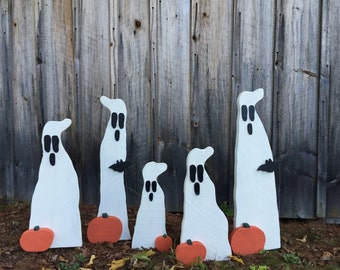 May include: Five white wooden ghost figures with black eyes and mouths, standing in a row in front of a wooden fence. Each ghost has a small orange pumpkin in front of it.