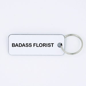 May include: White rectangular keyring with the text "BADASS FLORIST" in black lettering. The keyring has a silver metal ring.