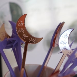 May include: A set of six purple and rose gold stirrers with moon-shaped toppers. The moon toppers are engraved with the text "Stay Wild Moon Child".