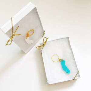 May include: Two white gift boxes with gold ribbon bows. Each box contains a gold key chain with a state-shaped charm. The charm in the left box is white, and the charm in the right box is turquoise blue.