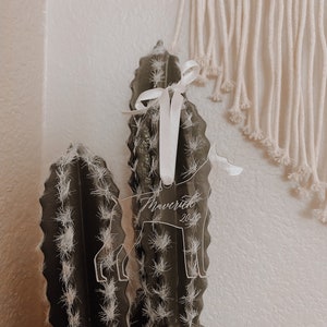 May include: A clear acrylic ornament in the shape of a dog, with the name "Maverick" and the year "2020" etched on it. The ornament is tied with a white ribbon and is displayed on a cactus.