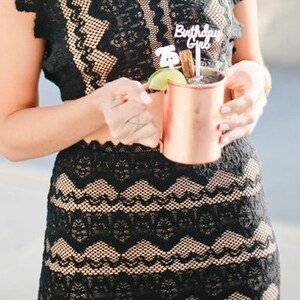 May include: A person wearing a black lace dress holds a copper mug with a lime wedge and a cocktail stirrer. The stirrer has a small sign that says "Birthday Girl".