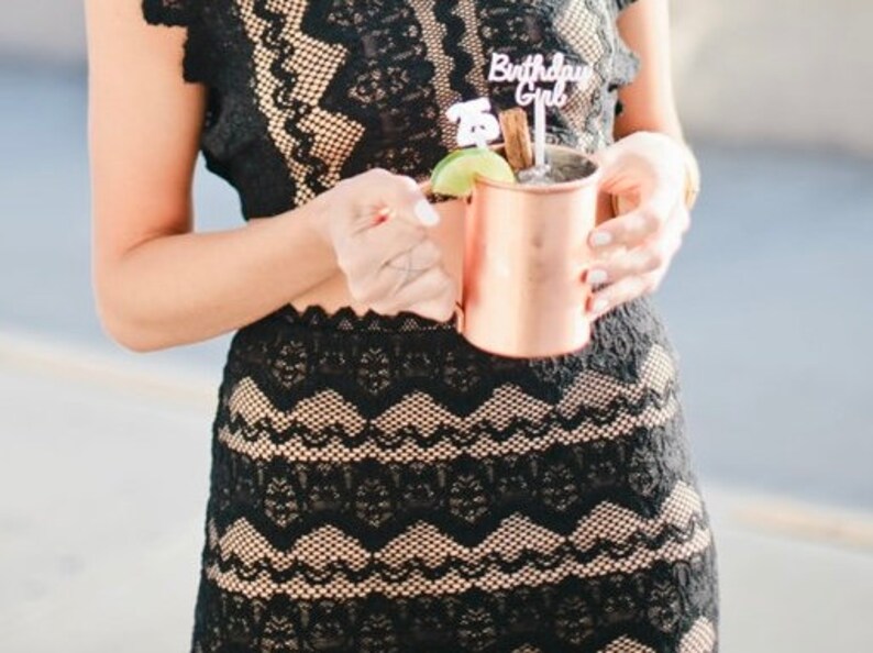 May include: A person wearing a black lace dress holds a copper mug with a lime wedge and a cocktail stirrer. The stirrer has a small sign that says "Birthday Girl".