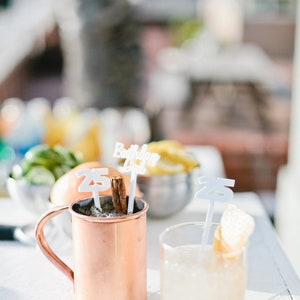 May include: Two copper mugs with drinks and a cinnamon stick. One mug has a white plastic sign that says "Birthday Girl" and a white plastic number "25". The other mug has a white plastic number "25" and a slice of lemon.