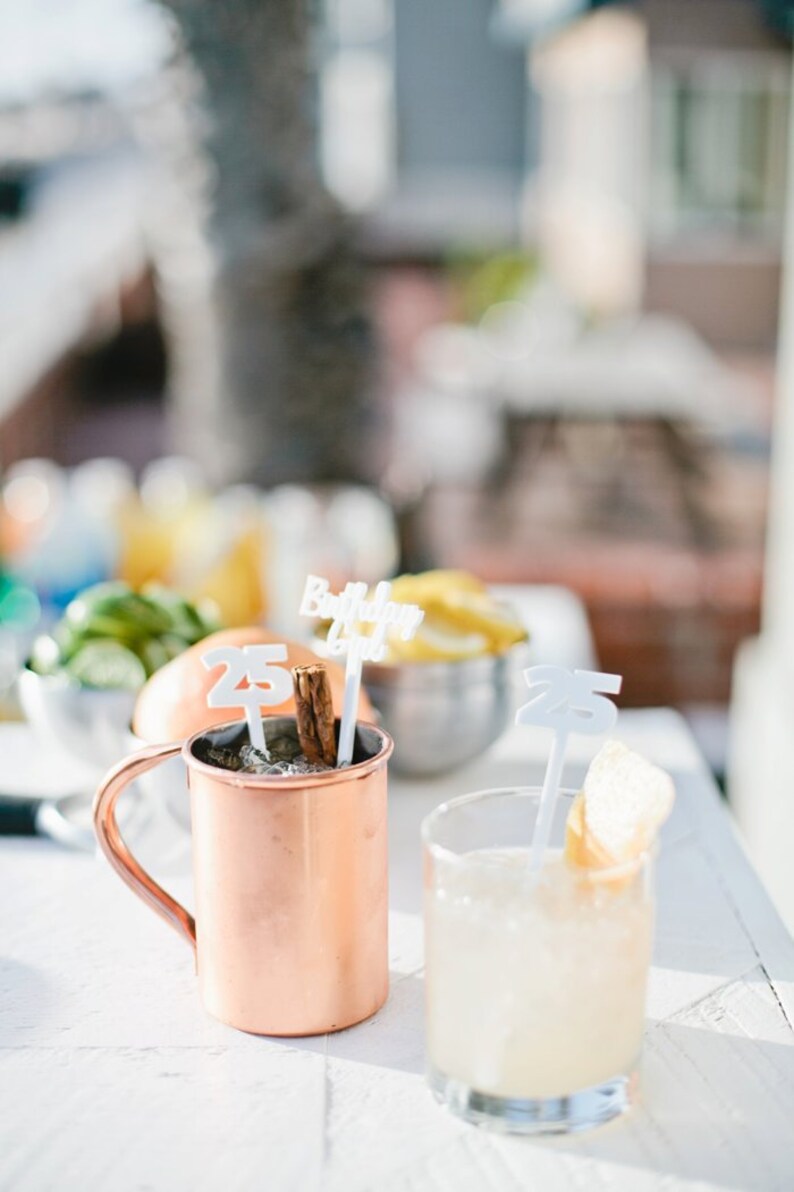May include: Two copper mugs with drinks and a cinnamon stick. One mug has a white plastic sign that says "Birthday Girl" and a white plastic number "25". The other mug has a white plastic number "25" and a slice of lemon.