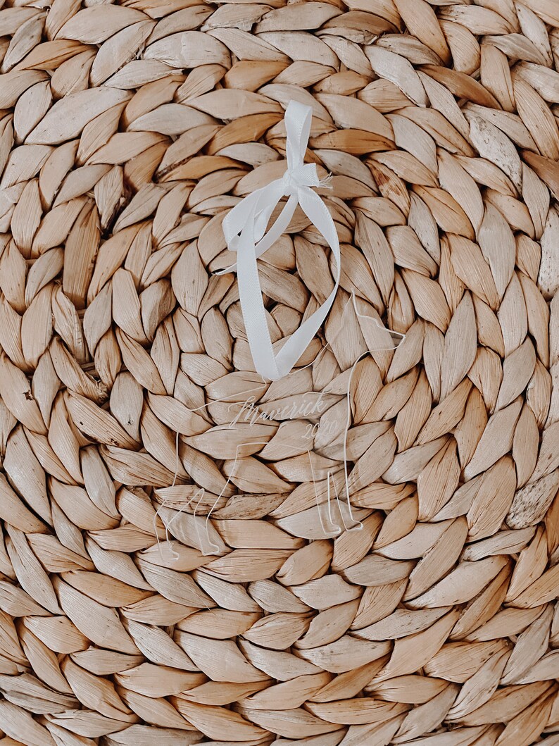 May include: A clear acrylic ornament in the shape of a dog, with the name "Maverick" etched on it. The ornament hangs from a white ribbon against a woven, light brown background.