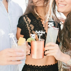 May include: Three people are holding cocktails with decorative 25th birthday themed stirrers. One cocktail is in a copper mug, one is in a clear glass with a lime wedge, and the other is in a tall glass with a cherry.