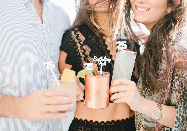 May include: Three people are holding cocktails with decorative 25th birthday themed stirrers. One cocktail is in a copper mug, one is in a clear glass with a lime wedge, and the other is in a tall glass with a cherry.