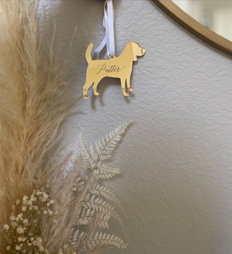 May include: A gold-colored dog-shaped ornament with the name "Putter" in cursive script. The ornament hangs from a white ribbon. The background includes dried plants and a wall.