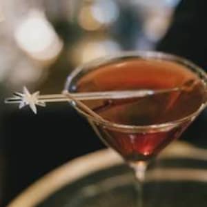 May include: A clear cocktail stirrer with a star-shaped topper resting across the rim of a martini glass filled with a dark amber liquid. The stirrer is transparent and features a multi-pointed star design at one end, extending over the glass.