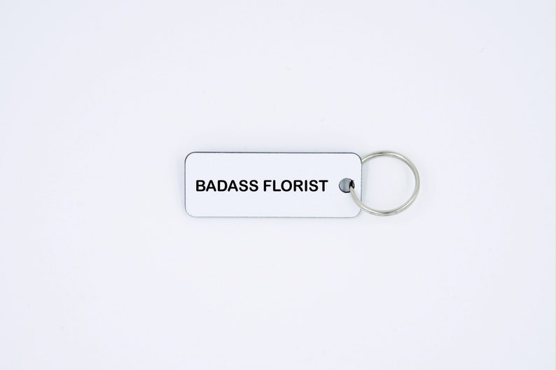 May include: White rectangular keyring with the text "BADASS FLORIST" in black lettering. The keyring has a silver metal ring.