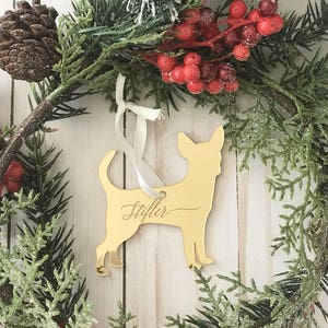 May include: A gold-colored Chihuahua dog ornament with the name "Stiffler" in cursive script, hanging from a white ribbon. The ornament is set against a Christmas wreath with red berries, pine needles, and a pine cone.