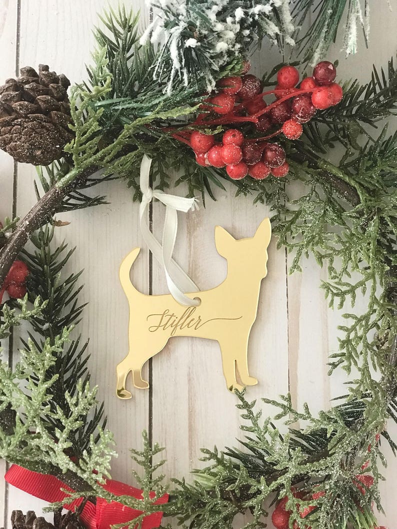 May include: A gold-colored Chihuahua dog ornament with the name "Stiffler" in cursive script, hanging from a white ribbon. The ornament is set against a Christmas wreath with red berries, pine needles, and a pine cone.