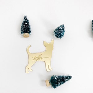 May include: A gold-colored Chihuahua ornament with the name "Stifler" engraved on it. The ornament is surrounded by small, green Christmas trees with white snow-like accents, all set against a white background.