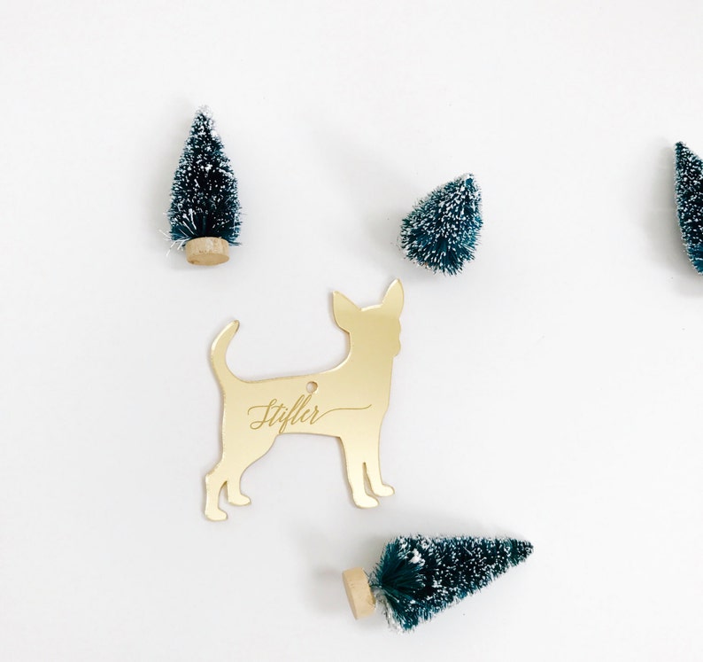 May include: A gold-colored Chihuahua ornament with the name "Stifler" engraved on it. The ornament is surrounded by small, green Christmas trees with white snow-like accents, all set against a white background.