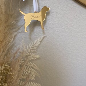 May include: A gold-colored dog-shaped ornament with the name "Beau" engraved on it. The ornament hangs from a white ribbon. The background includes dried flowers and a neutral-colored wall.