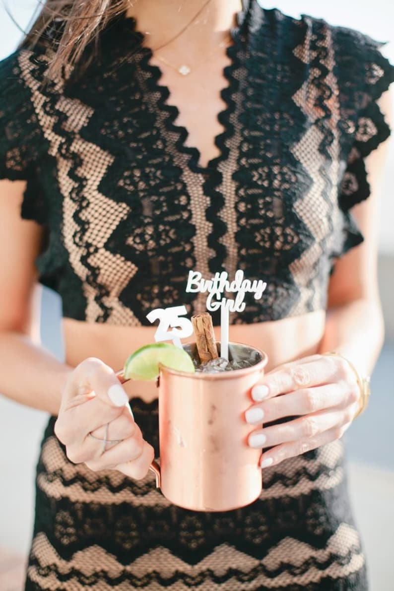 May include: A person holding a copper mug with a lime wedge and a cinnamon stick. The mug has a white paper straw with the words "Birthday Girl" and "25" on it.