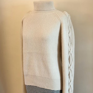 May include: A cream-colored turtleneck sweater with a ribbed knit and cable knit detailing on the sleeves.