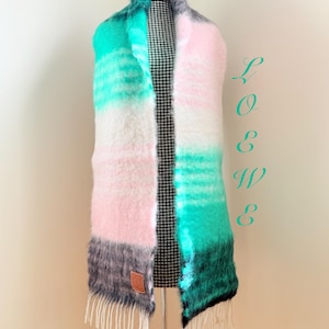 May include: A colorful scarf with a gradient design, featuring shades of pink, white, teal, and gray. The scarf has a fringed edge and a small leather tag. The scarf is displayed on a mannequin.