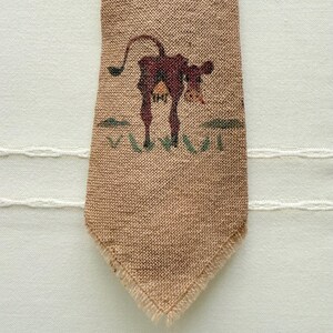 May include: A brown, textured fabric tie with a detailed illustration of a brown cow. The cow is standing on green grass with two green bushes on either side. The tie has a frayed edge.