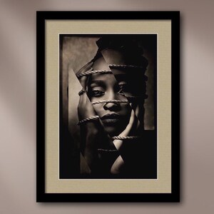 May include: A black and white photo of a person's face with rope wrapped around their head and hands. The photo is framed in a black frame with a beige mat.