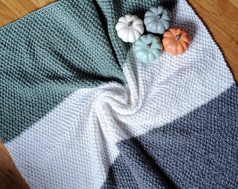 May include: A handmade, textured blanket in shades of green, white, and gray. The blanket is folded with decorative mini pumpkins in white, gray, teal, and orange. The blanket is made of a soft material and is perfect for home decor.