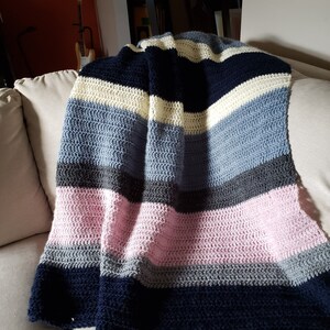 May include: A striped crocheted throw blanket in shades of navy blue, light blue, gray, pink, and cream. The blanket is folded and draped over a couch.