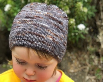 May include: A young child wearing a gray and brown striped knit hat is smelling a pink flower. The child is wearing a yellow long-sleeved shirt.