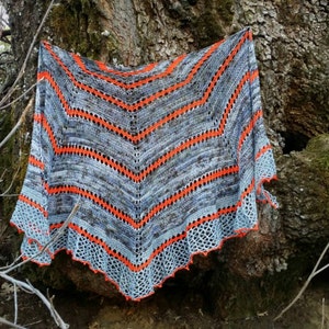 Puede incluir: Un chal de punto gris y naranja con un borde de encaje cuelga de un tronco de árbol.