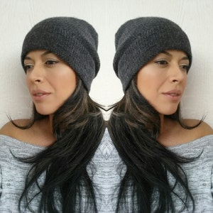 Puede incluir: Una mujer con un gorro de punto gris. El gorro es holgado y le cubre las orejas. Tiene el pelo largo y oscuro y lleva un jersey gris.