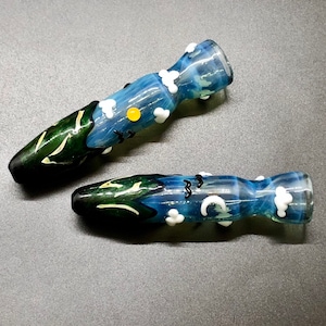 May include: Two glass pipes with a nature-inspired design. One pipe features a green and white leaf pattern, while the other has a blue sky design with white clouds, a yellow sun, and black birds. The pipes are on a gray surface.